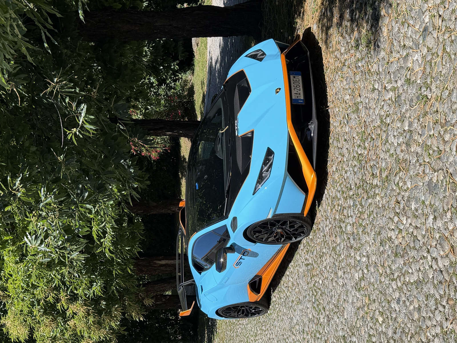 Lamborghini Huracán STO azzurra con dettagli arancio fotografata di tre quarti anteriore su un vialetto alberato.