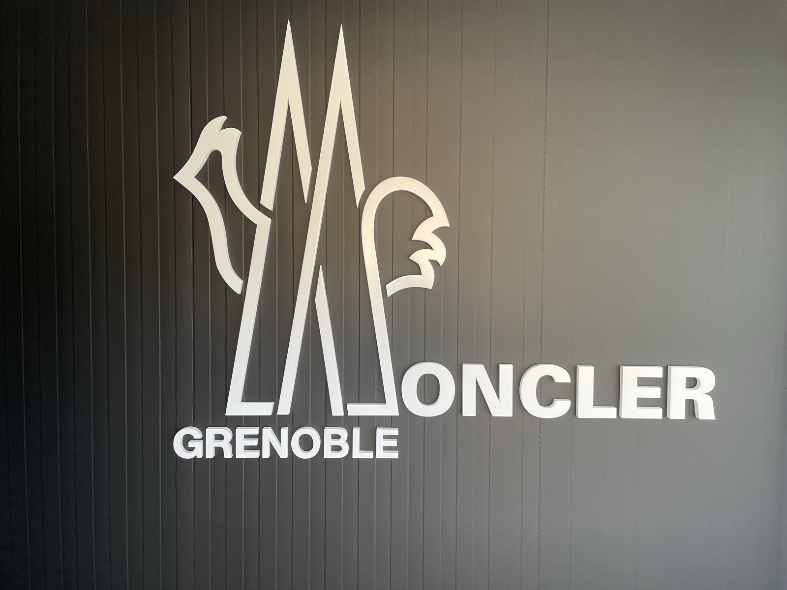 Grande logo bianco Moncler Grenoble applicato su una parete scura a righe verticali.