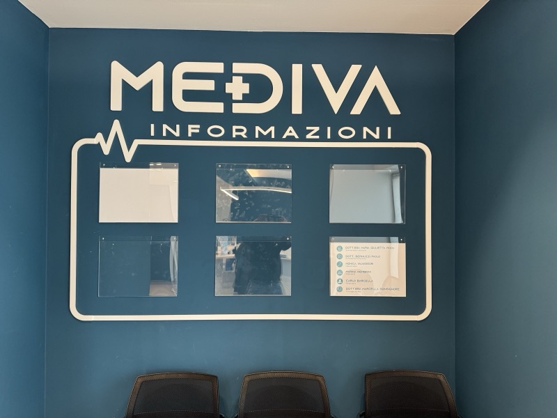 Bacheca informazioni Mediva bianca su una parete blu, con logo e spazi per avvisi.