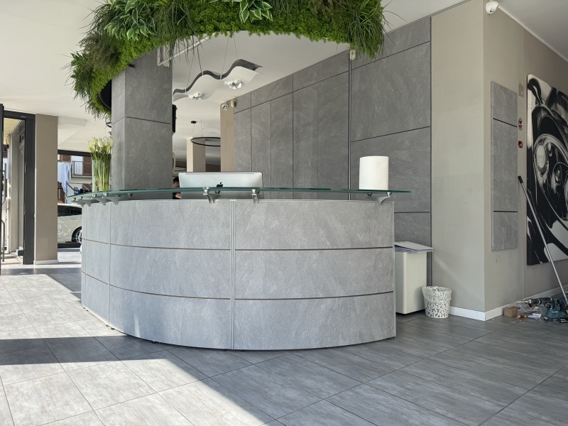 Bancone curvo e parete della reception con rivestimento grigio effetto pietra, specchio e piante sospese.