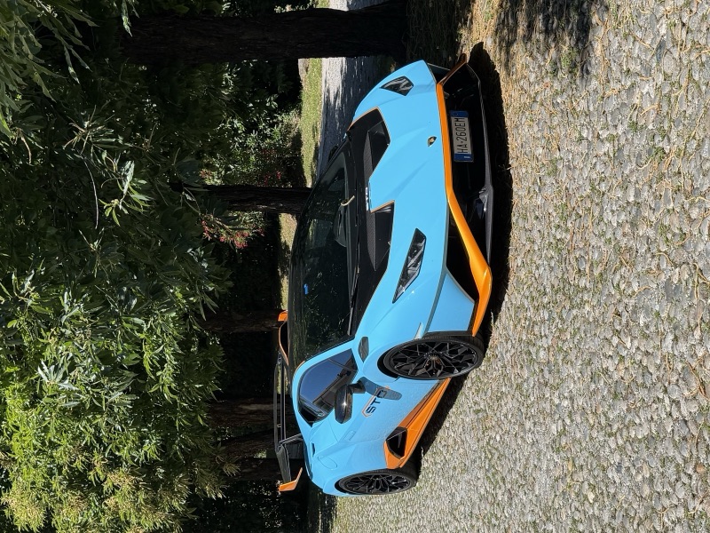 Lamborghini Huracán STO azzurra con dettagli arancio fotografata di tre quarti anteriore su un vialetto alberato.