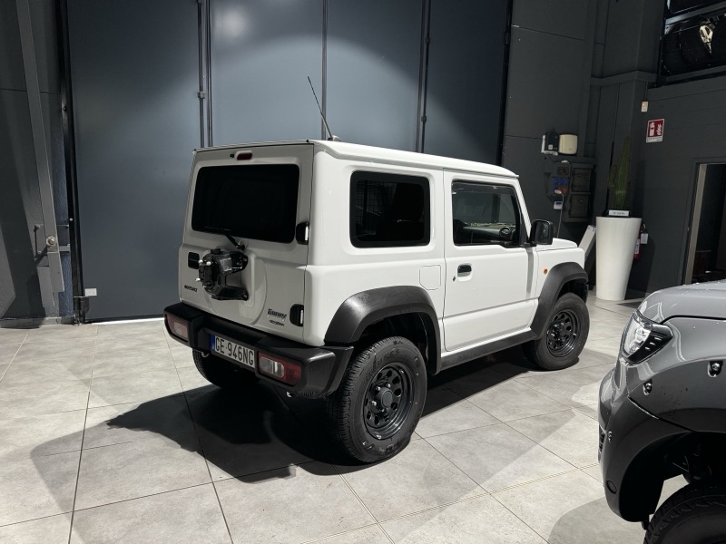 Suzuki Jimny bianco visto di tre quarti posteriore in officina, con lunotto e vetri posteriori oscurati.