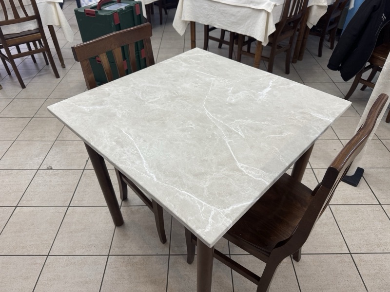 Piano quadrato di un tavolo da ristorante con finitura chiara effetto pietra, circondato da sedie in legno.